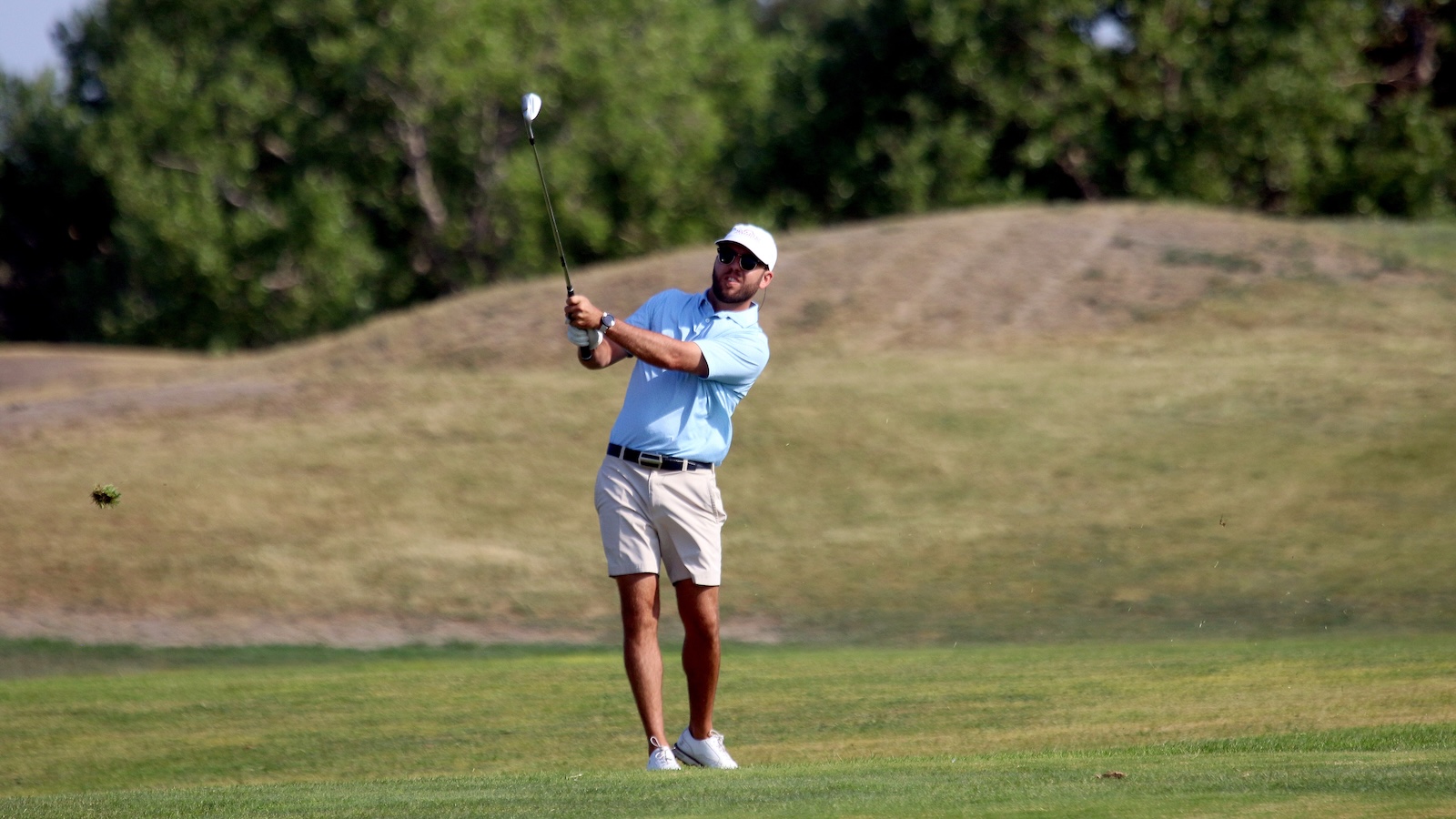 Golfer in a light blue polo and white cap mid-swing on a grassy golf course.