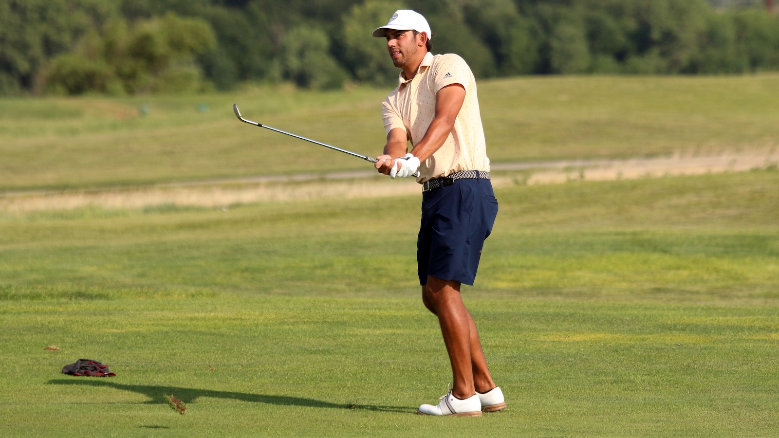 Man in a beige polo and navy shorts swinging a golf club on a green fairway; white cap, white shoes, glove on left hand, buggy item on ground nearby