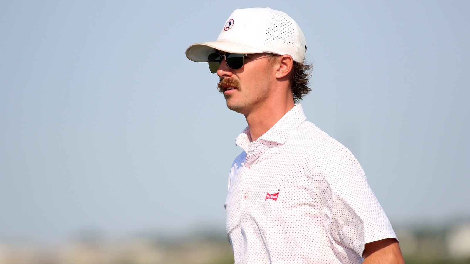 Male golfer in a white cap and sunglasses, looking to the right against a clear blue sky.