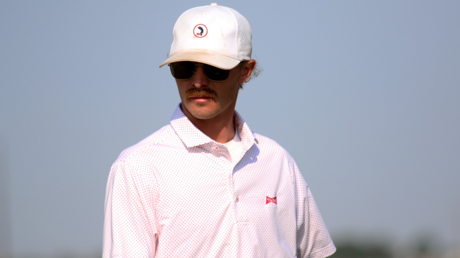 Man wearing a white cap, sunglasses, and a pale pink dotted polo shirt outdoors with a mustache
