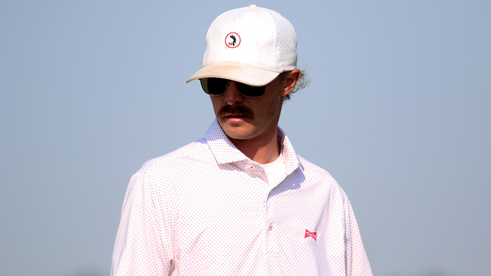Man with a white cap, dark sunglasses, and a light pink dotted shirt outdoors on a sunny day, looking to the side.