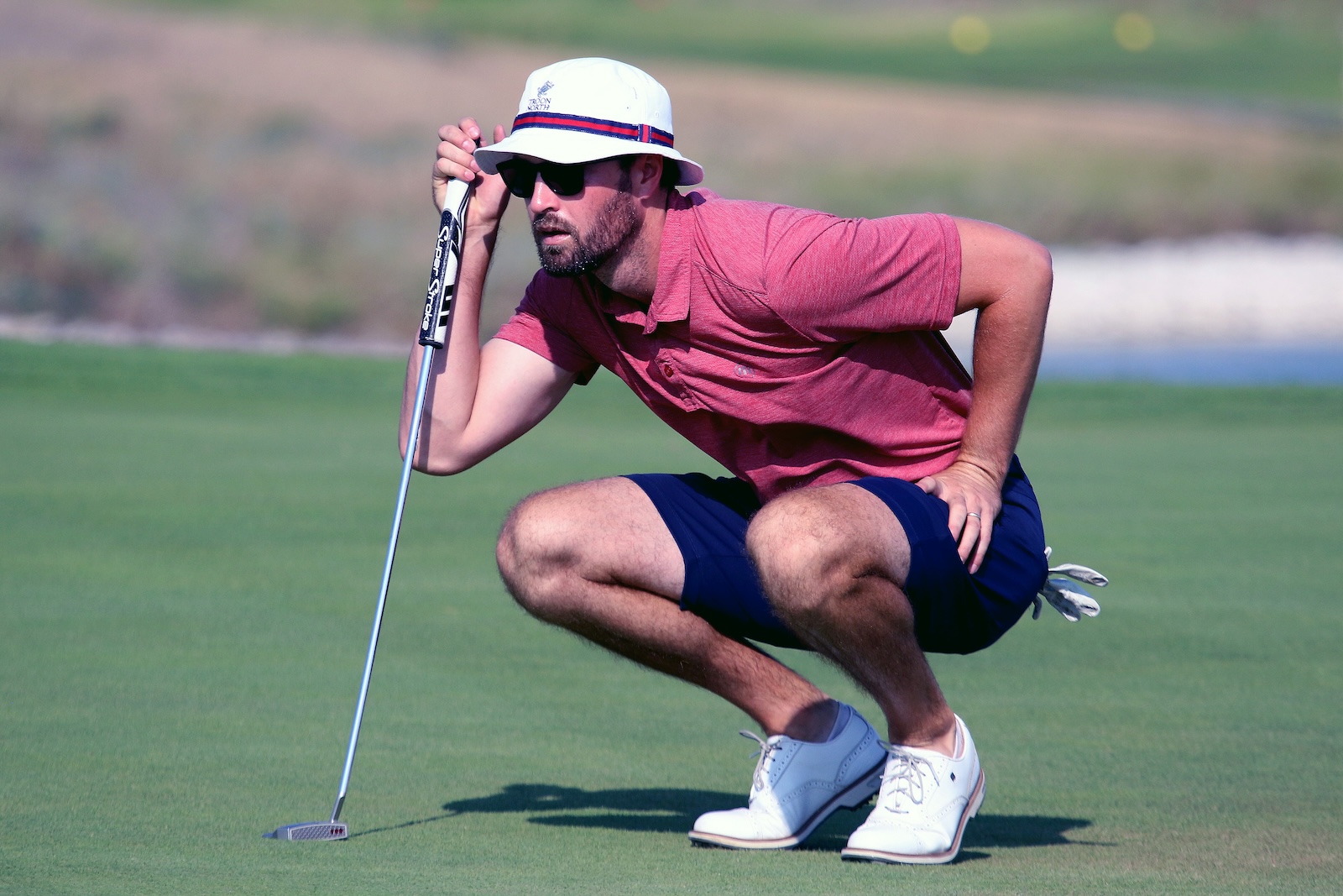 Male golfer crouches on the green, lining up a putt with a white putter, wearing a white hat, sunglasses, red polo, and navy shorts.