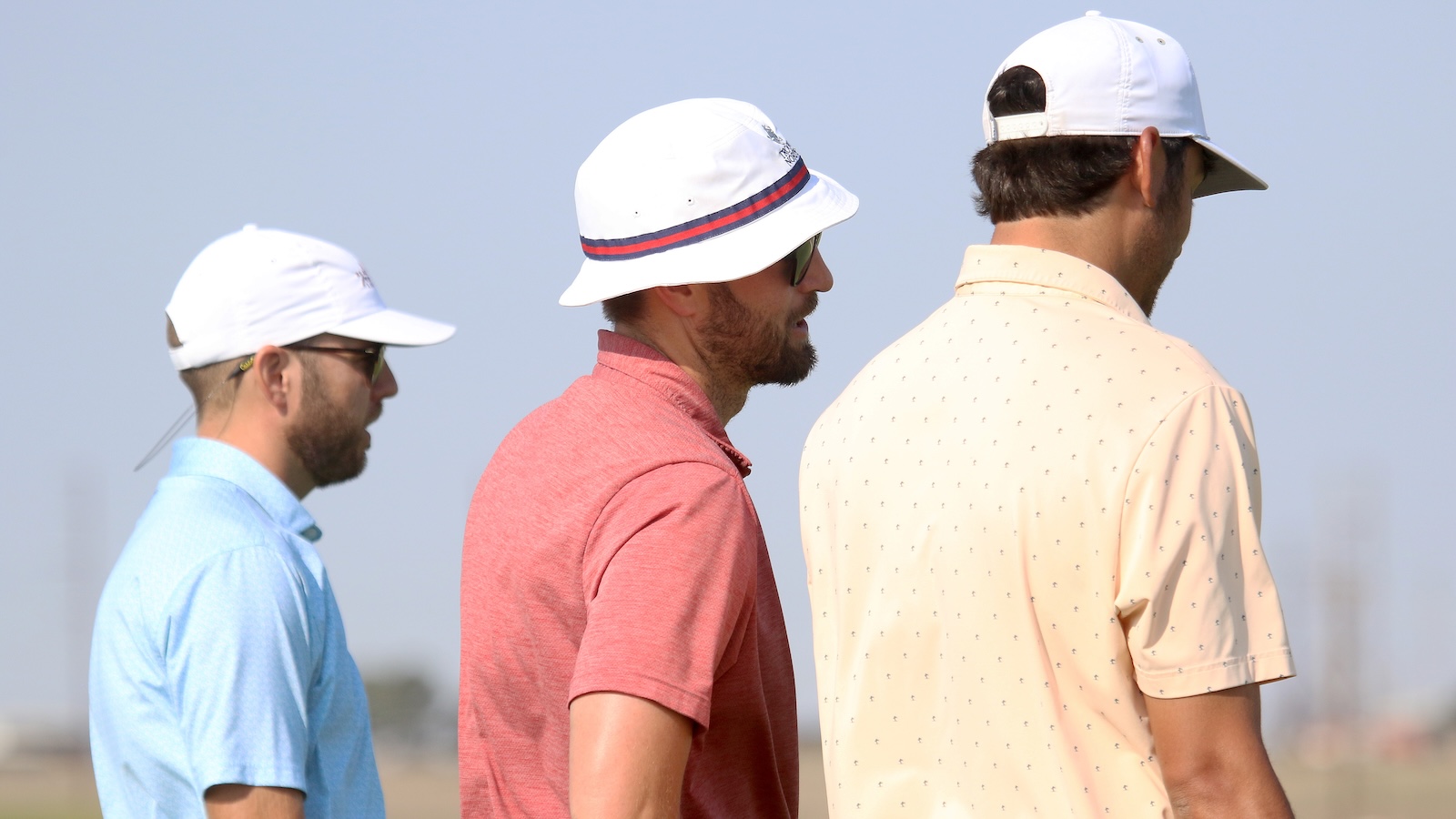 Three men in light blue, coral, and pale peach polo shirts wear white caps and stand side by side outdoors on a sunny day.