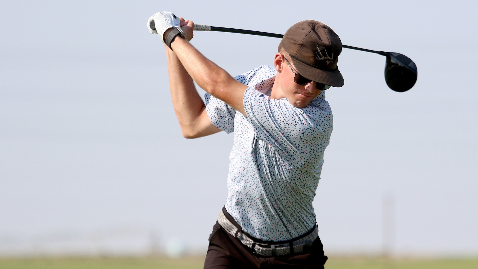 Golfer in a patterned shirt swinging a golf club with a brown cap and sunglasses on a sunny day