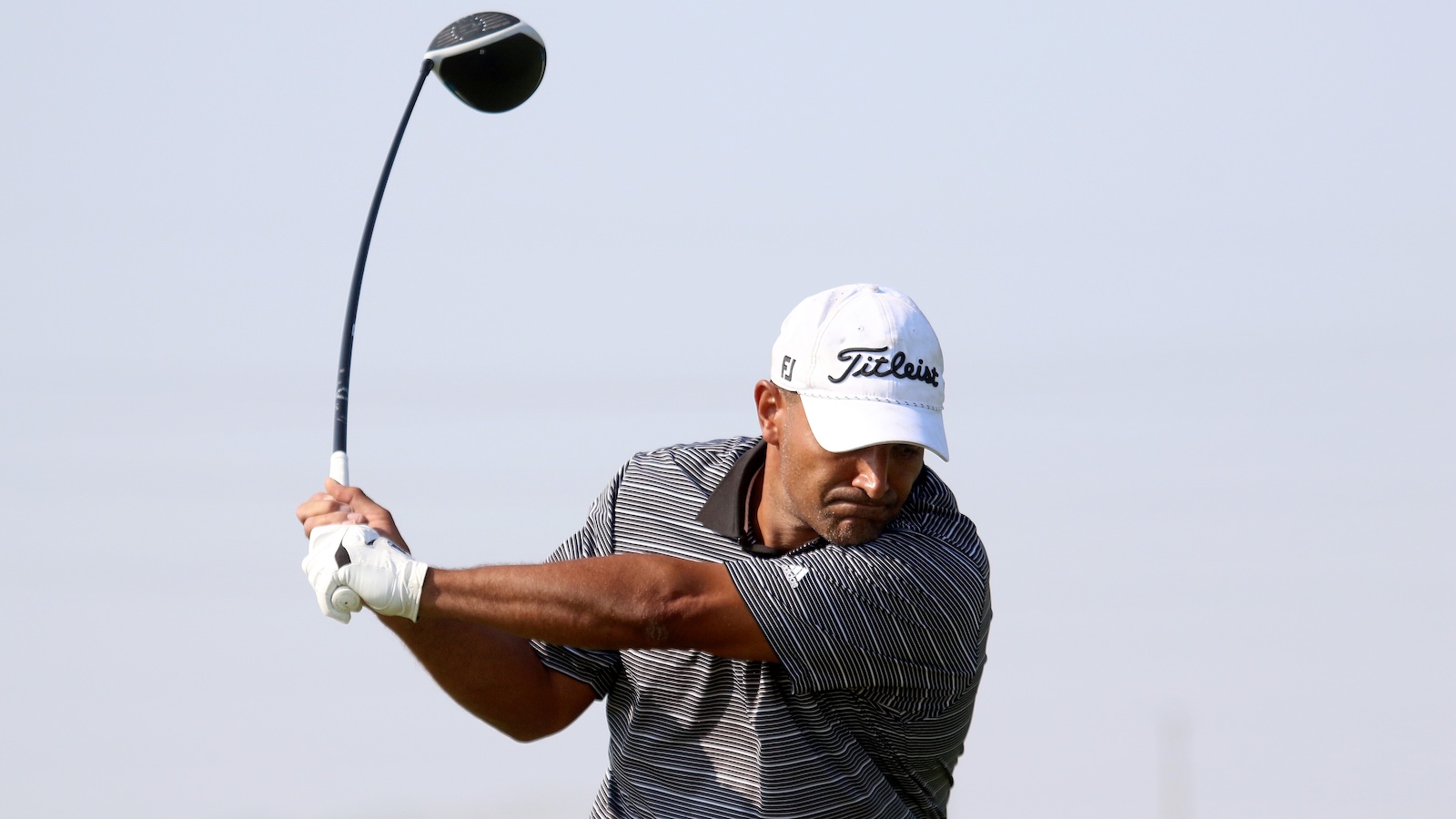 Male golfer in a striped shirt and white Titleist cap swinging a golf club.