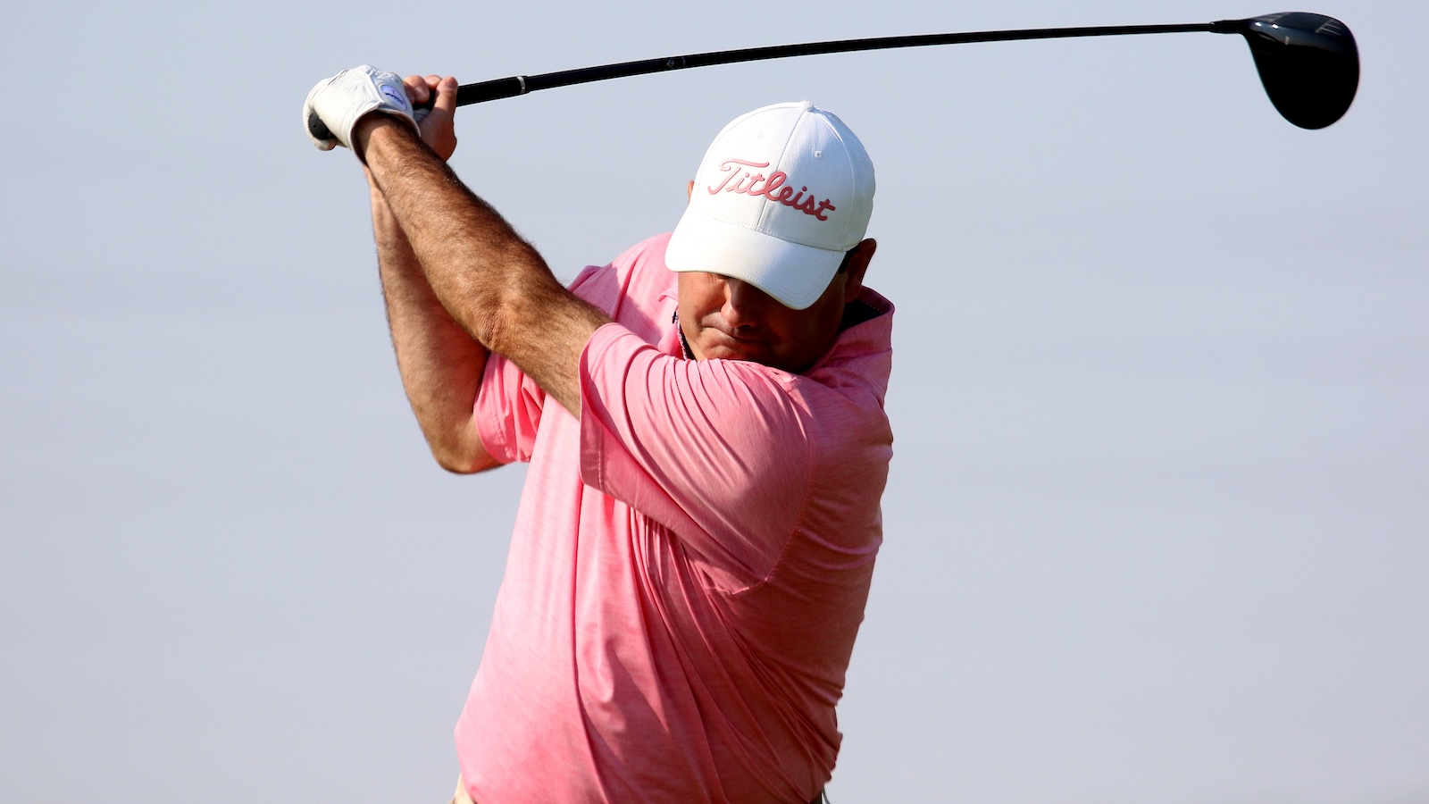 Male golfer in a pink shirt swinging a golf club, wearing a white Titleist cap against a clear sky