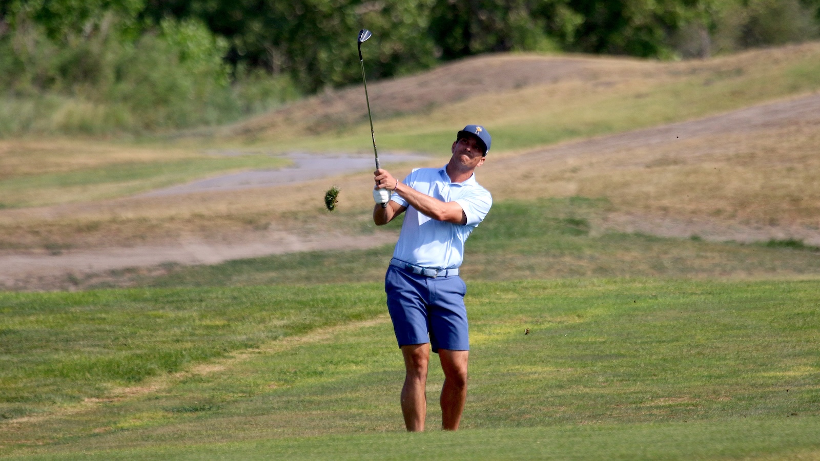 Man in a light blue polo and navy shorts mid-swing on a golf course, with green grass and trees in the background now following through on the drive.