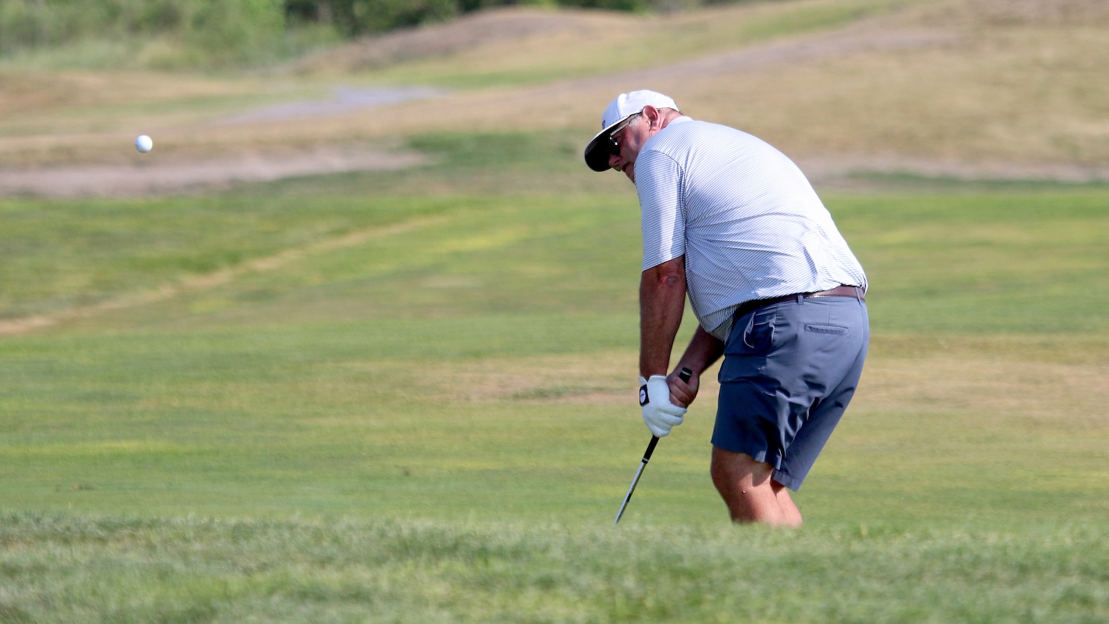 Golfer in a white cap and striped shirt swings at a golf ball on a sunlit course.