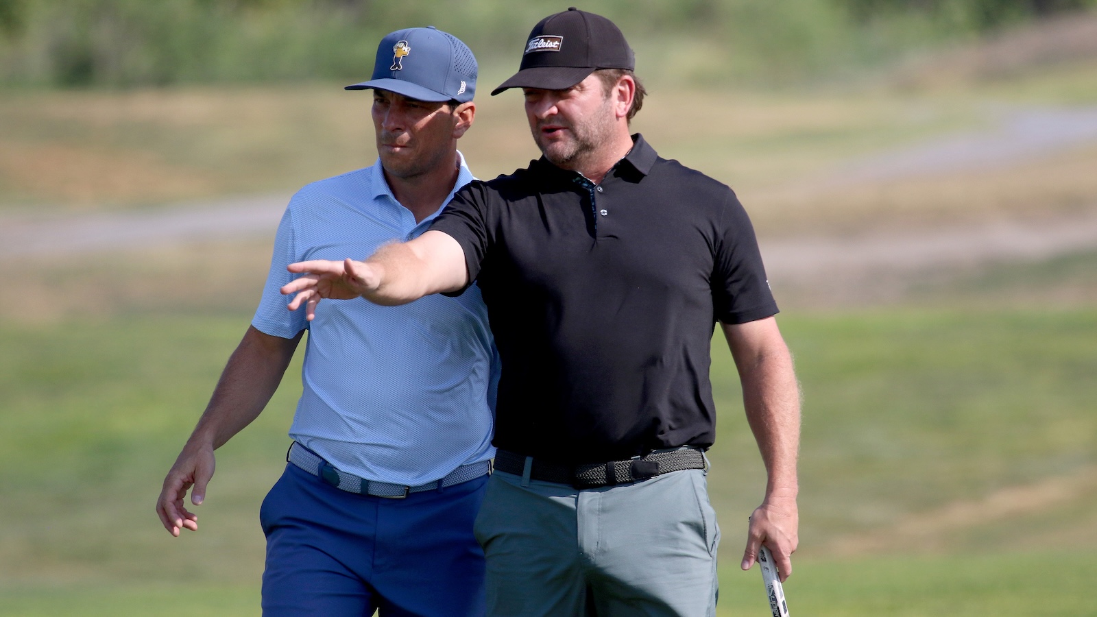 Two men on a golf course: one in a black polo points ahead while the other in a light blue shirt walks beside him.