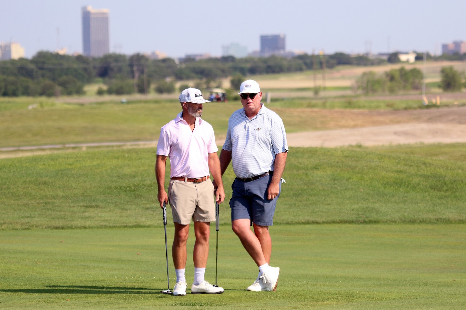 Two golfers standing on a green, chatting with putters in hand, city skyline in the distance.