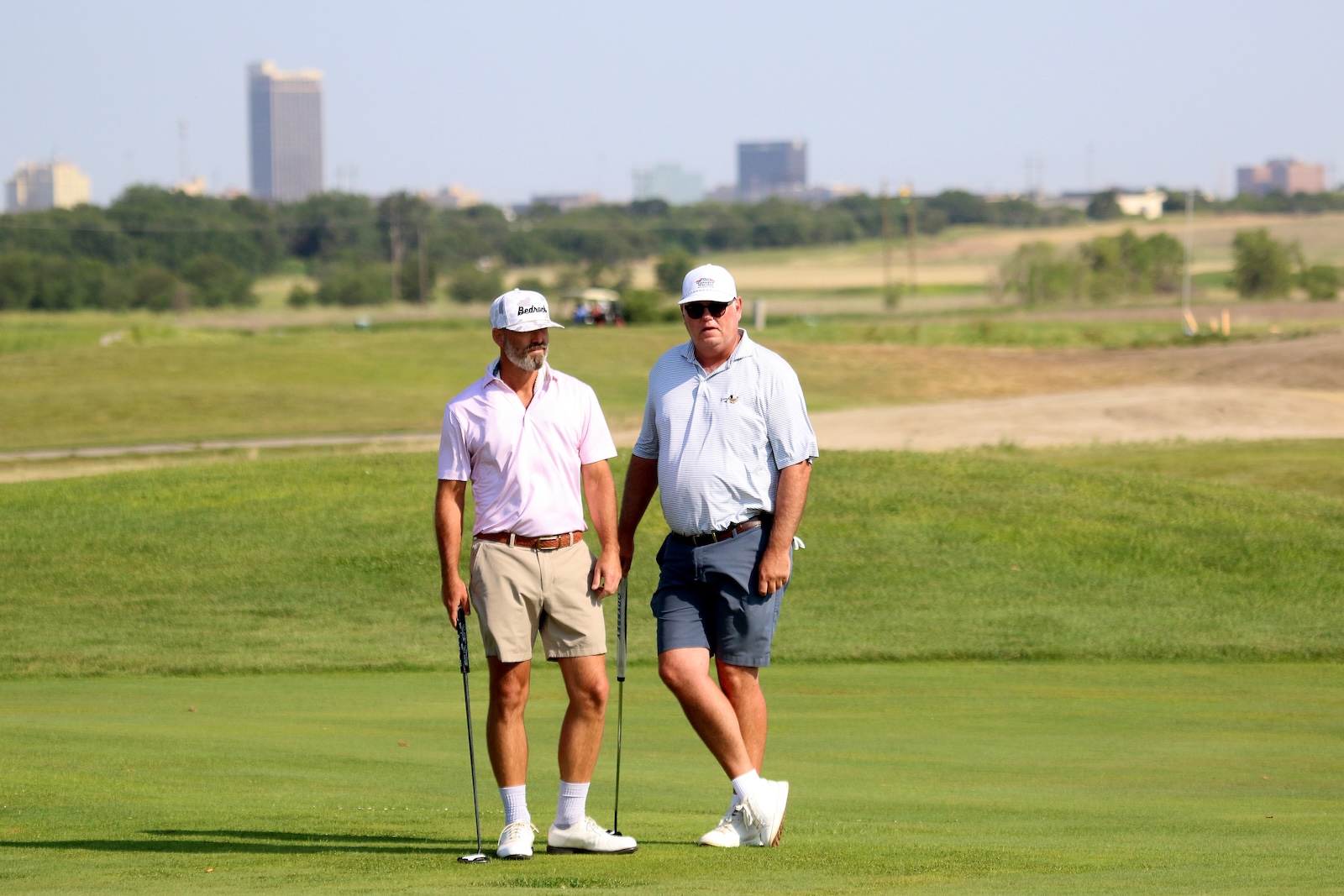 Two men in polo shirts and caps walk with putters on a golf green, city skyline in distance.