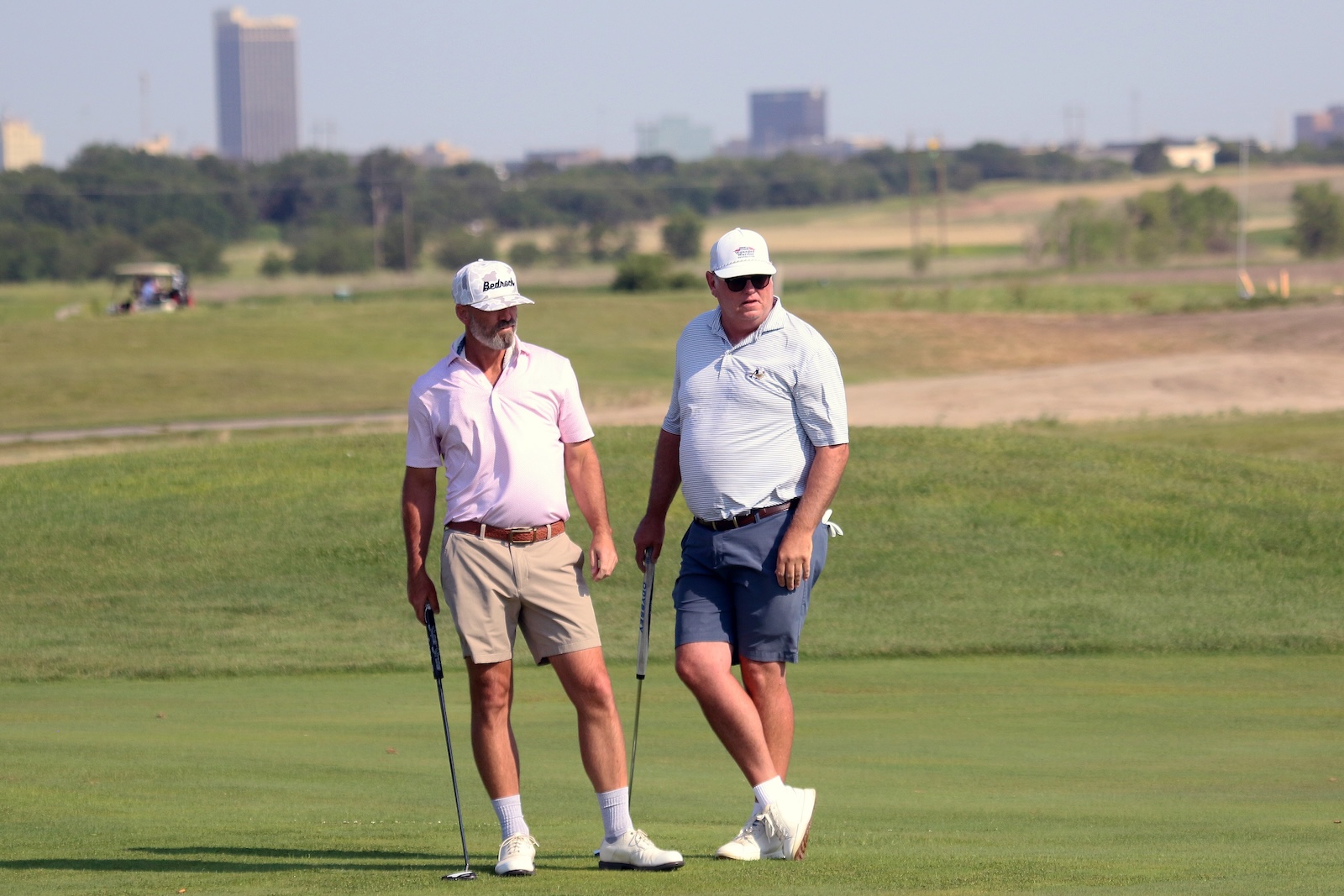 Two men in polo shirts and caps stand on a golf course with a city skyline in the background.