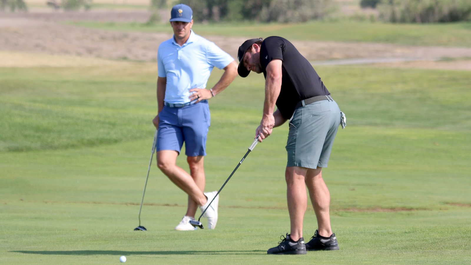 Two men on a golf course; one is putting while the other watches nearby.