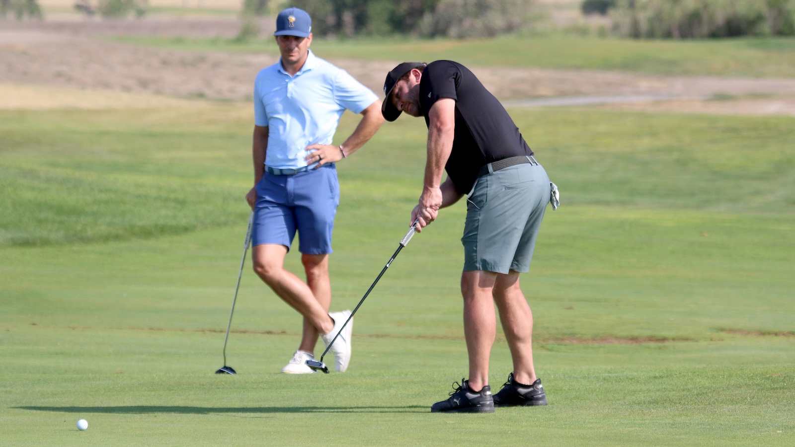 Two golfers on the green: one in a black shirt lines up a putt while a teammate in light blue watches nearby with hands on hips.