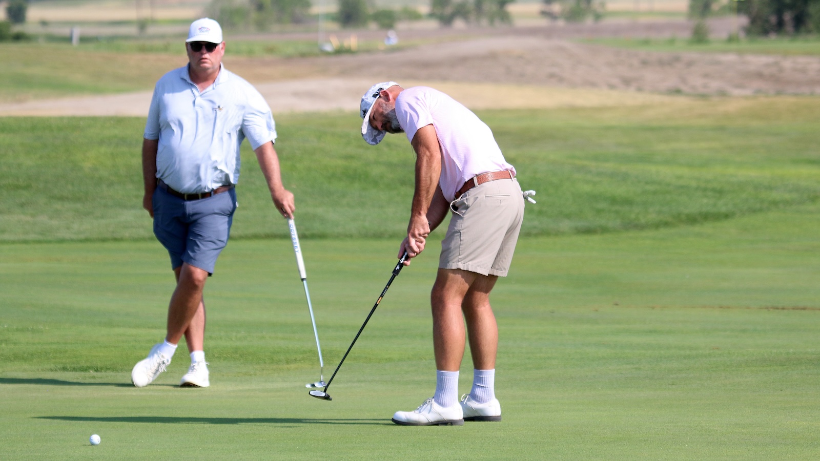 Two golfers on a sunny golf course: one bending to putt, the other standing and watching nearby.