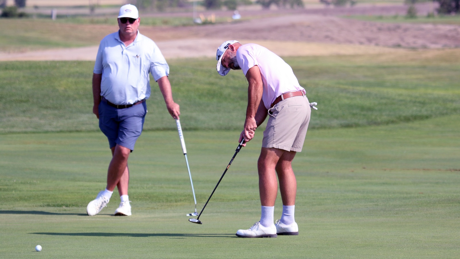 Two golfers on a green: a man putting while another man watches nearby in light polo and shorts.