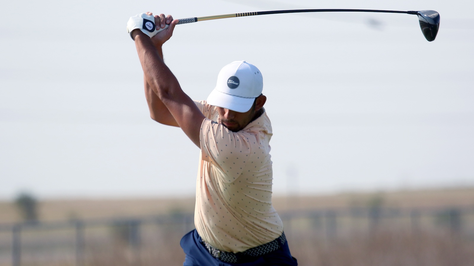 Golfer in a white Titleist cap and beige dotted polo mid-swing with a driver on a sunny course.