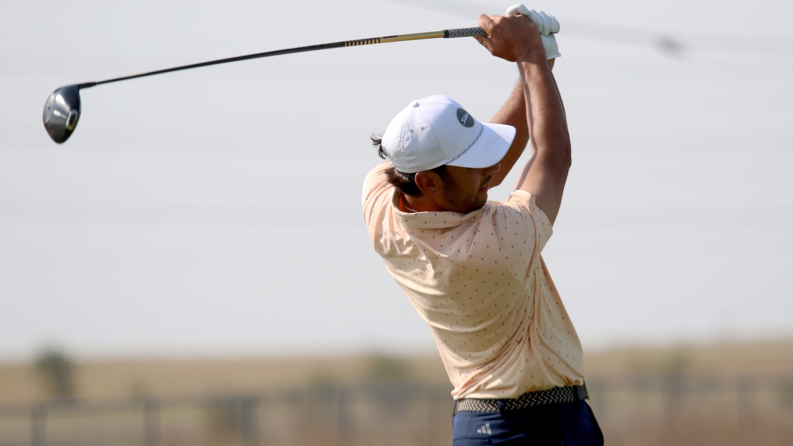 Golfer mid-swing on a sunny course, wearing a white cap and light peach polo.