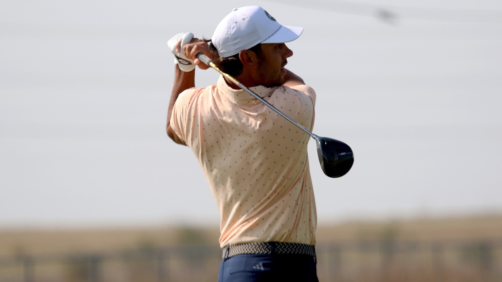 Golfer in peach polo and white cap mid-swing, backswing with driver on a sunny day
