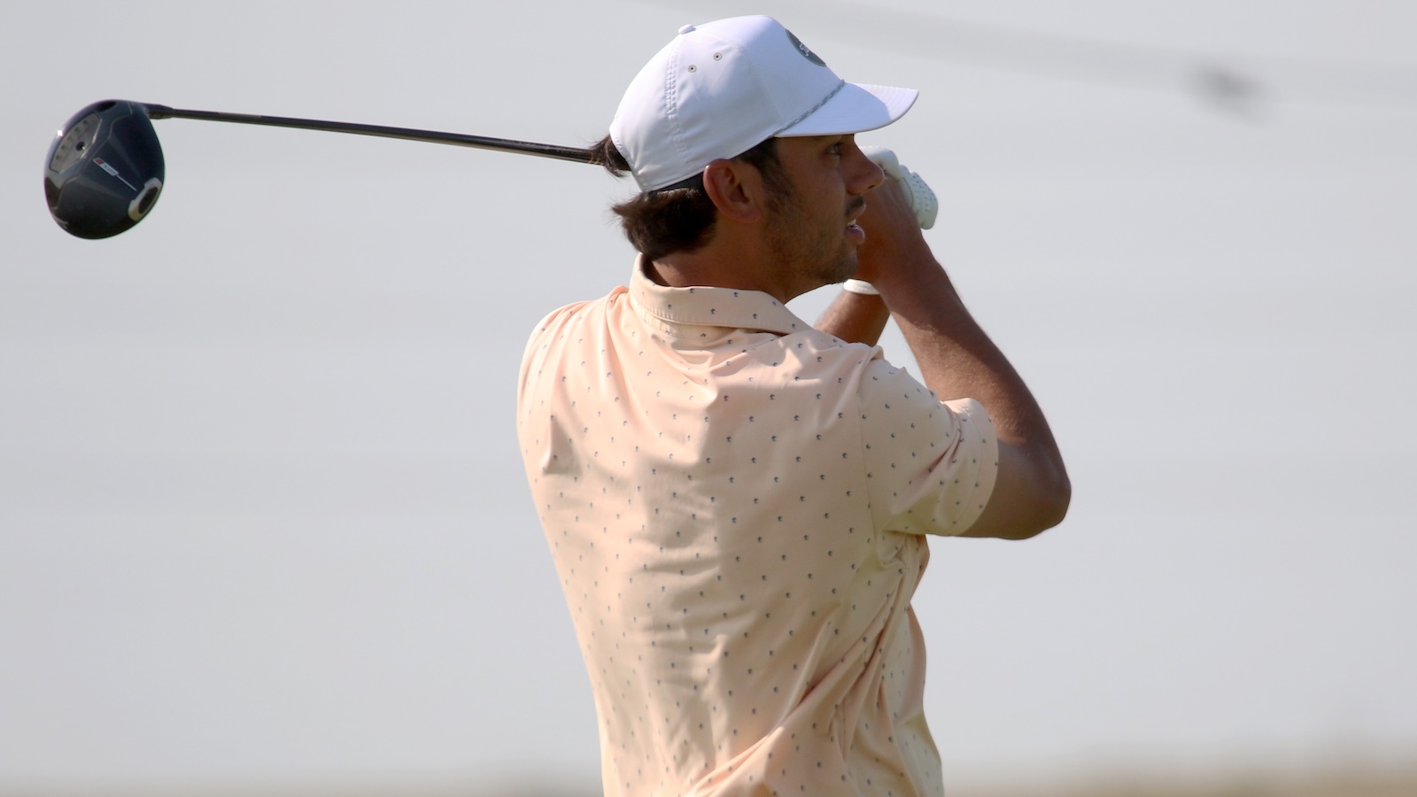 Golfer in a peach polo and white cap mid-backswing on a golf course.