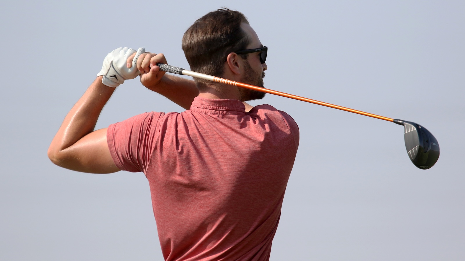 Man in a red shirt takes a backswing with a golf driver, seen from behind on a sunny day with a clear sky.