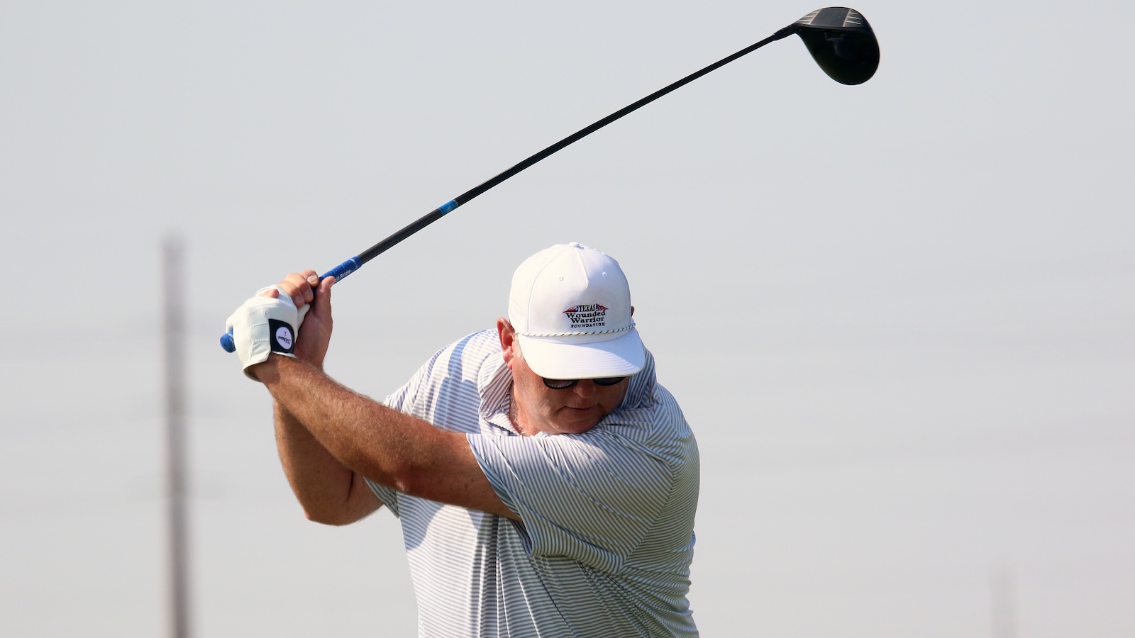 Man in a light striped polo swings a golf club during a drive on a sunny day.
