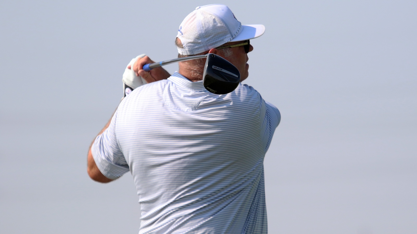 Male golfer in a white cap and striped polo swings a club in a backswing, facing away from the camera against a clear sky, sunglasses on.
