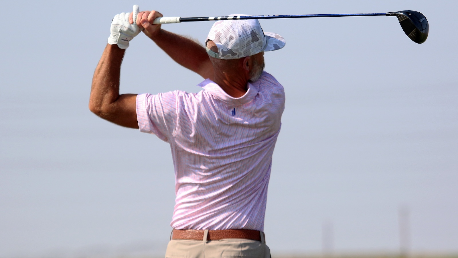 Male golfer in a pink polo and cap mid backswing with a golf club on a sunny course, seen from behind.