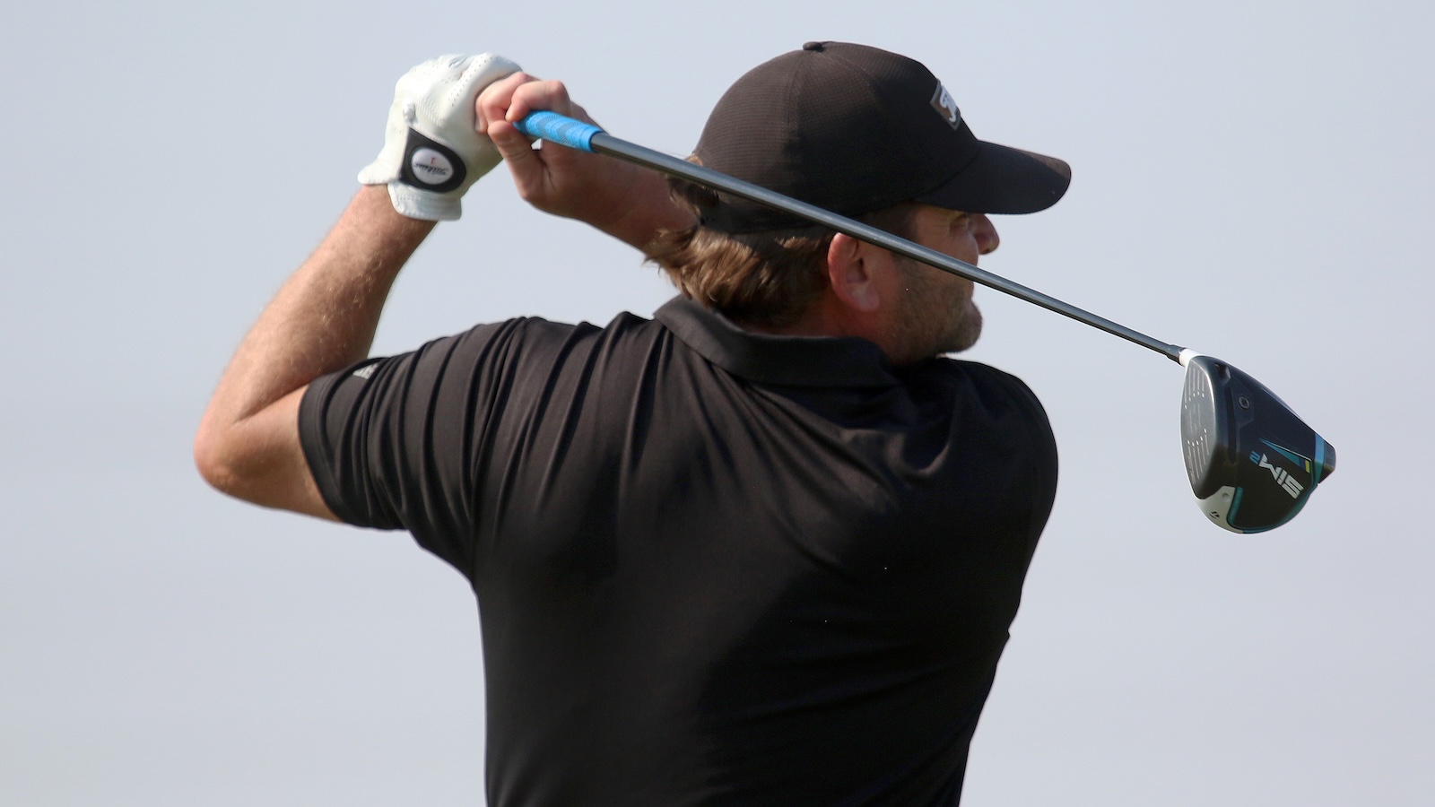 Golfer in a black shirt and cap mid-swing with a driver, wearing a white golf glove on the left hand, outdoors