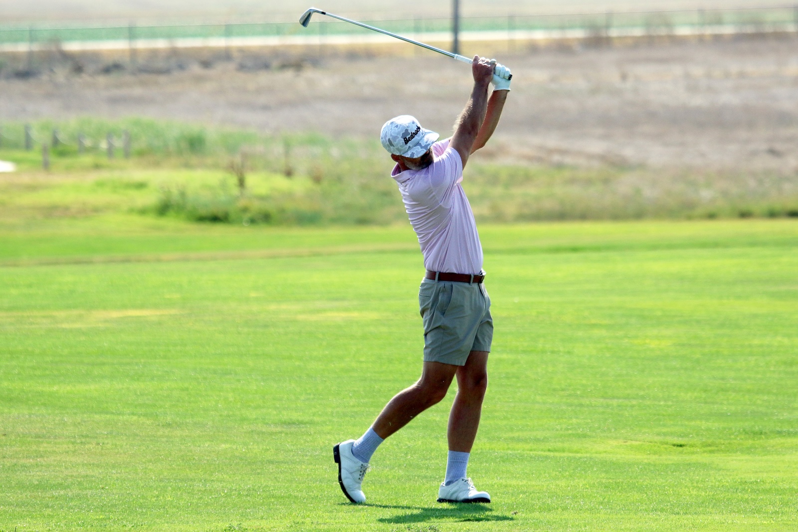 Male golfer mid-swing on a green golf course, wearing a light pink polo, gray shorts, white cap, and white shoes.