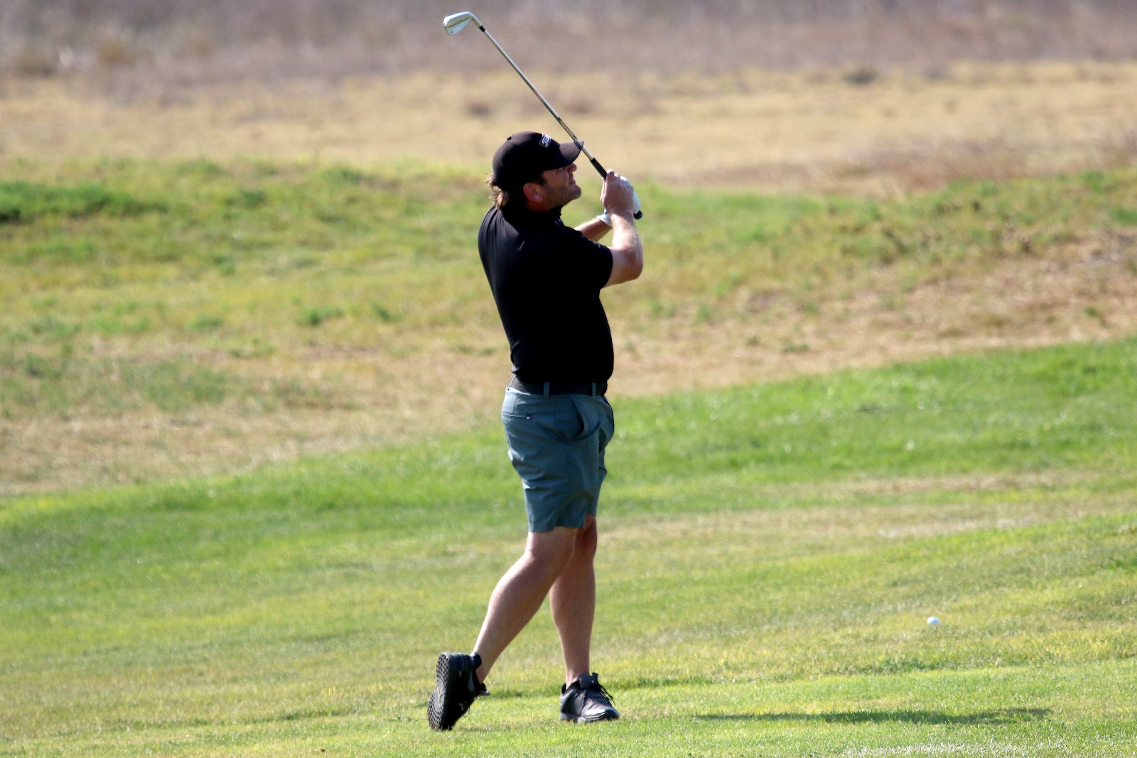 Man in a black polo and teal shorts swinging a golf club on a grassy golf course.