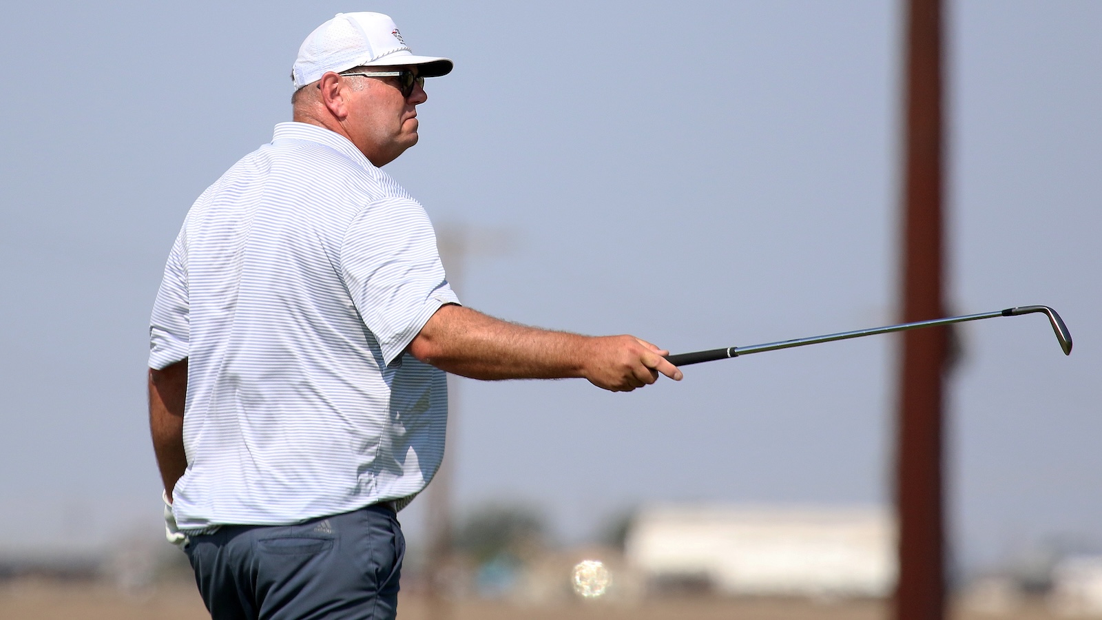 Middle-aged man wearing a white cap and striped polo, sunglasses, extending a golf club to the right on a course.