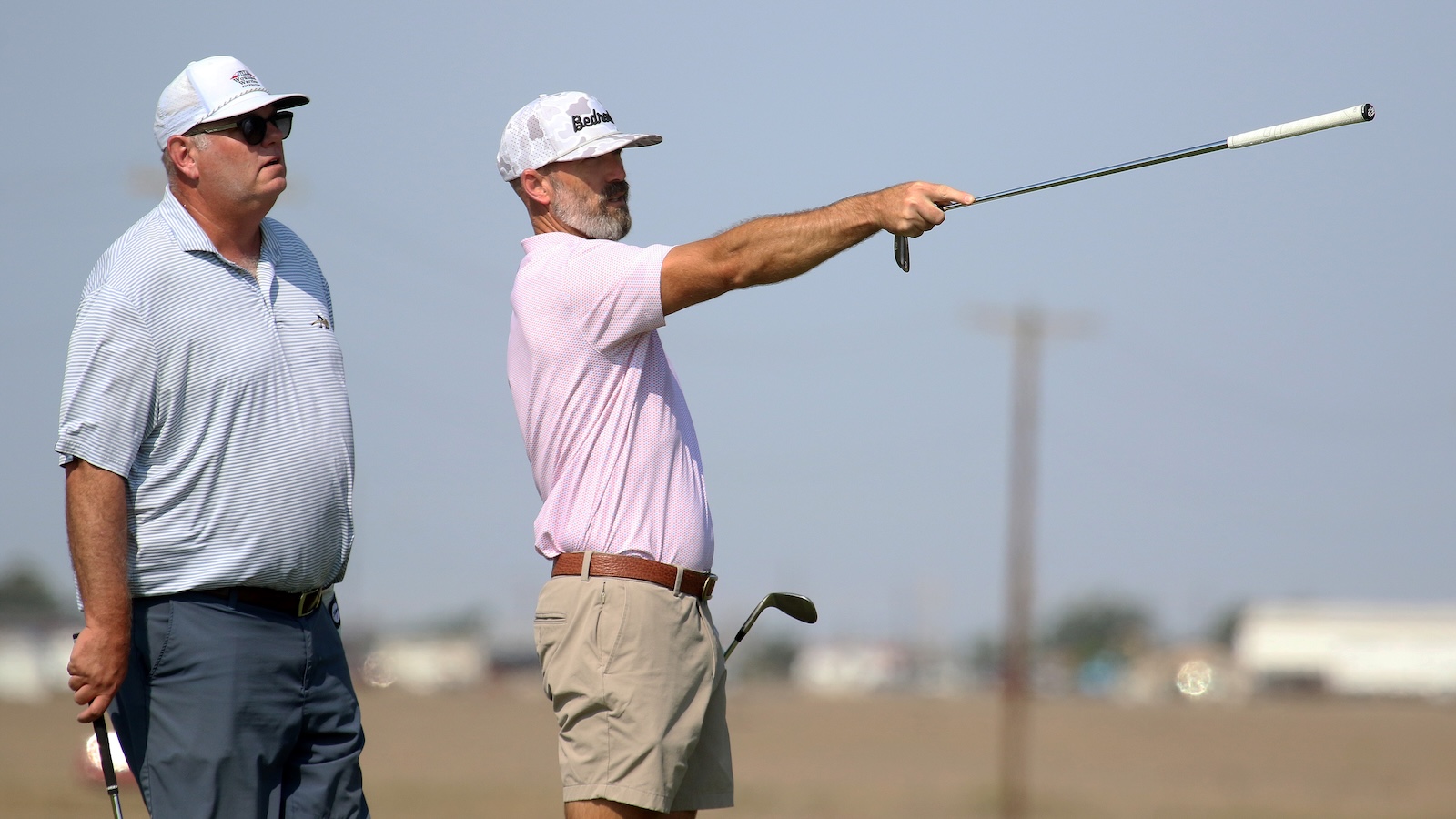 Two men on a golf course: one in a pink shirt swinging a club, the other watching nearby in a striped polo.