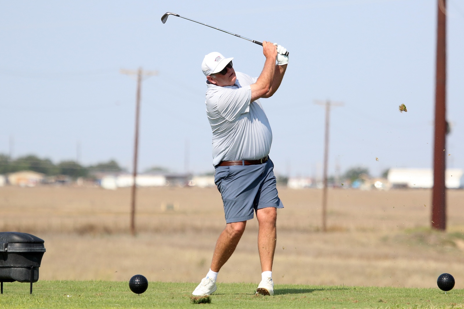 Man in a white cap, striped polo, and blue shorts swings a golf club on a sunny driving range; a golf ball sits on the tee near his feet.