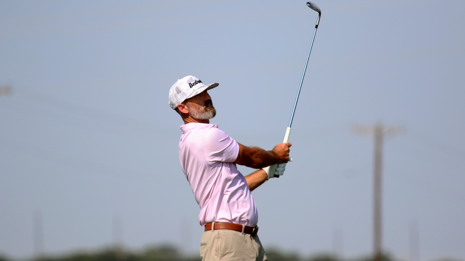 Older man in a pink polo and white cap mid-swing with a golf club on a sunny day on the course.