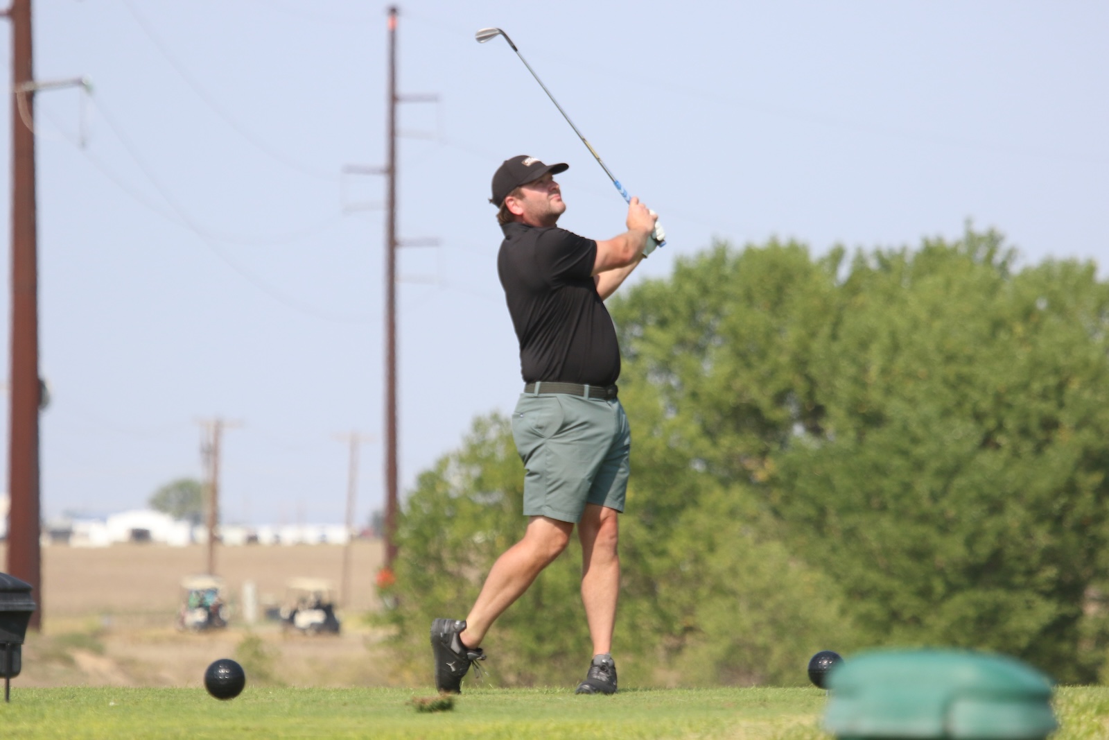 Male golfer in a black polo and gray shorts mid-swing on a sunny course, with golf balls on the grass nearby.