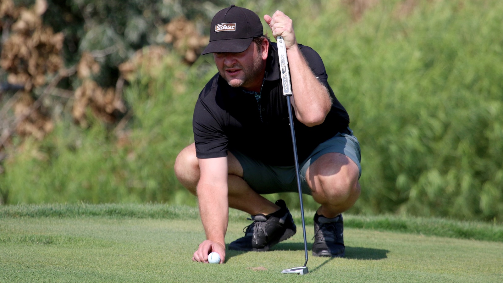 Golfer crouching on the green, lining up a putt with a putter and golf ball nearby under a sunny sky.