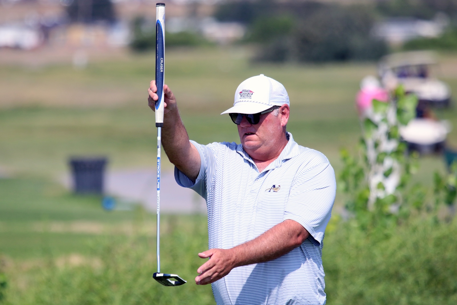 Man in a white cap and striped polo raises a putter on a sunny golf course.