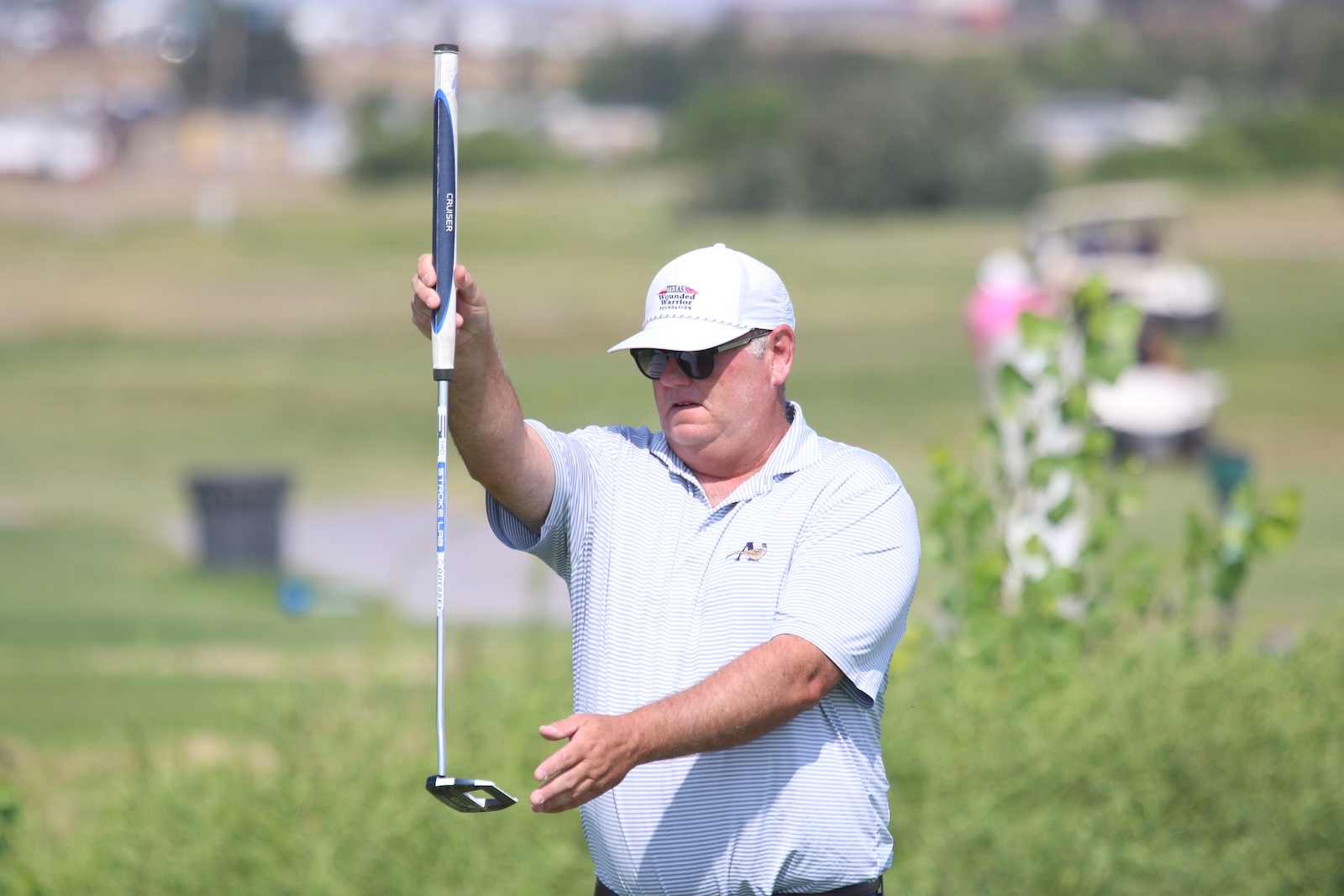 Male golfer in a light polo and white cap holds a putter upright on a sunny golf course.