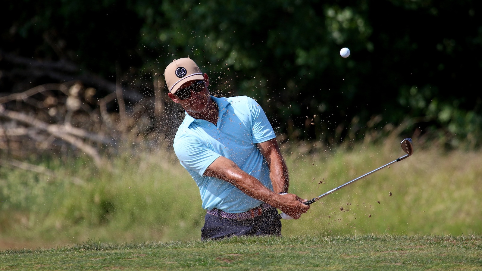 Golfer in a light blue polo and tan cap hits a sand shot from a bunker, ball in midair with sand spray.