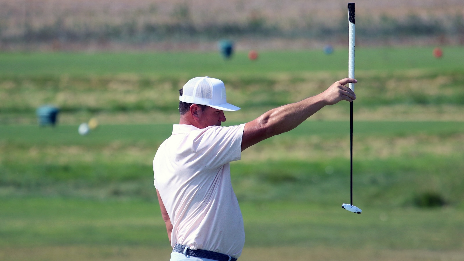 Man in a white polo and cap holds up a long golf alignment rod in a grassy practice field