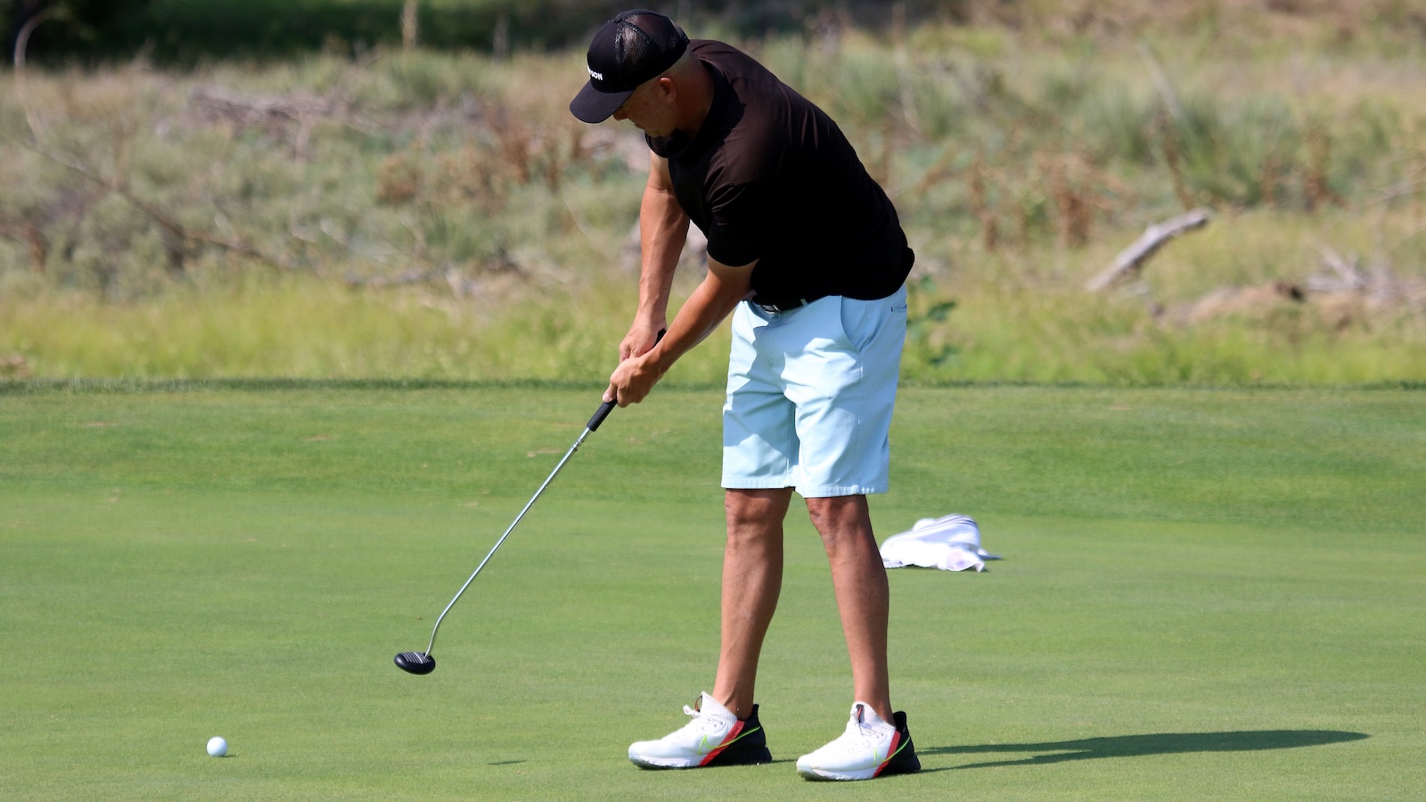 Golfer bending over to putt on a green, wearing a black shirt, light blue shorts, and white sneakers with a golf club ready to strike a ball.
