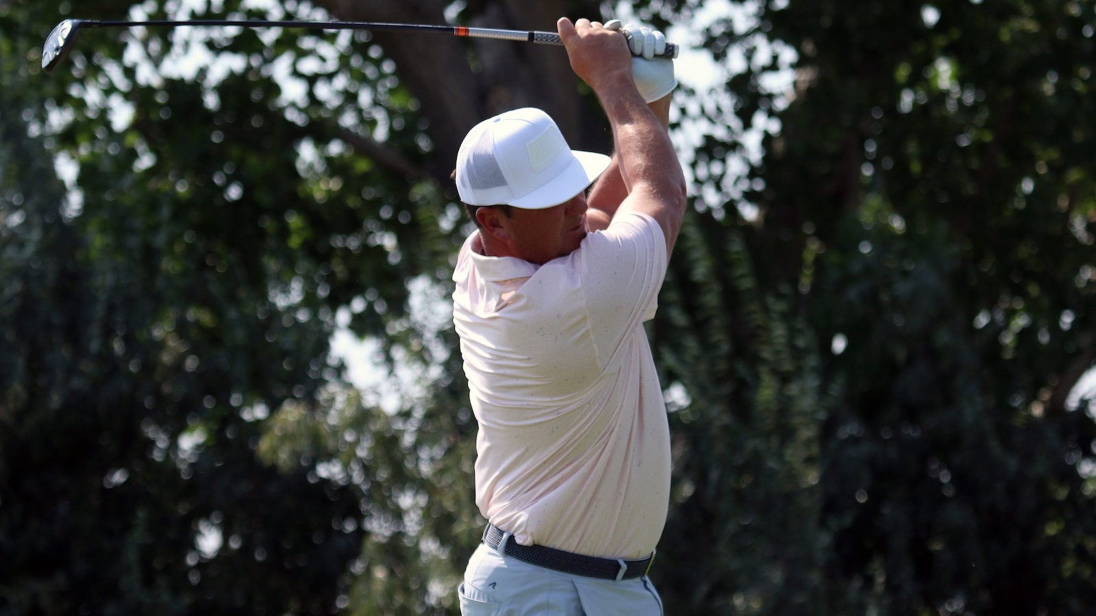 Golfer wearing a white cap and pale pink polo mid-swing with a driver on a sunny course.