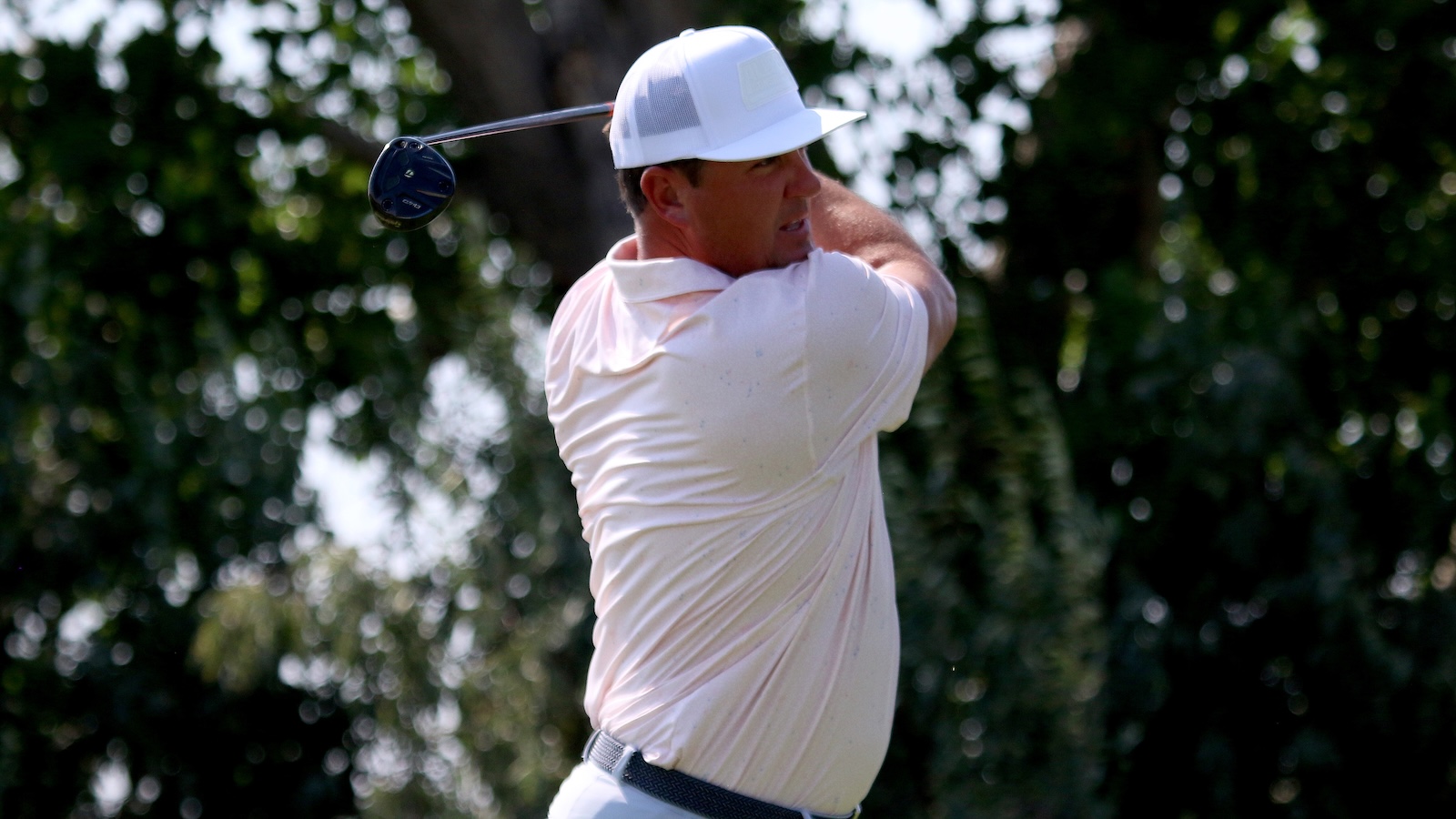 Man in a white polo and cap mid-swing on a golf course, preparing to hit the ball with a driver.
