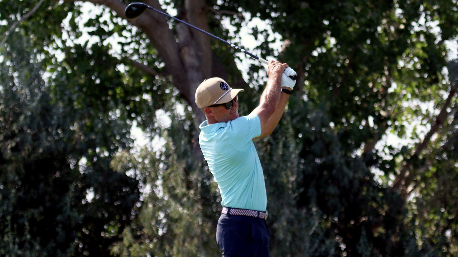Golfer in a light blue polo swings a club on a sunny day with trees in the background.