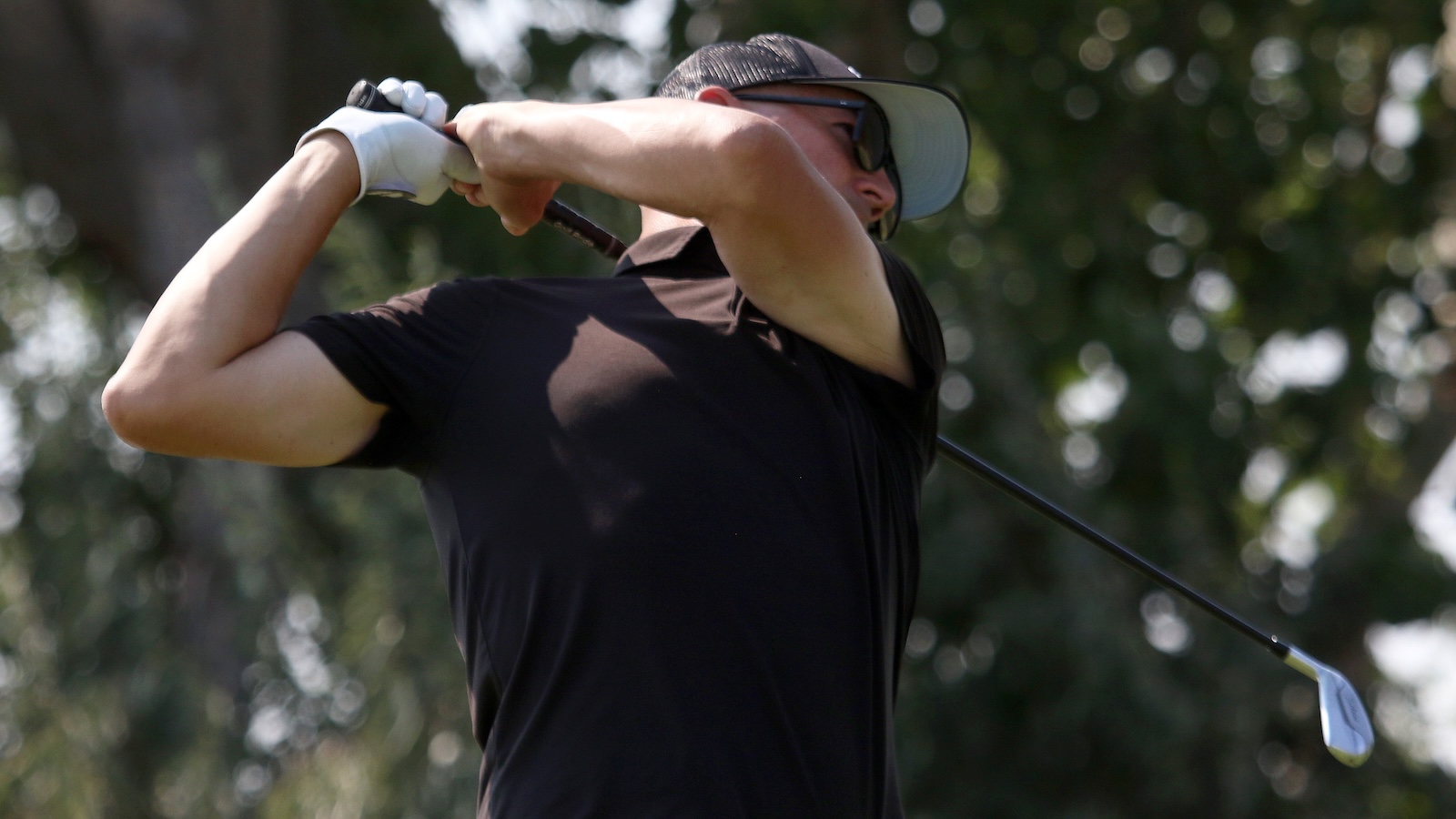 Golfer in a black shirt mid-swing on a sunny day, wearing a cap and sunglasses.