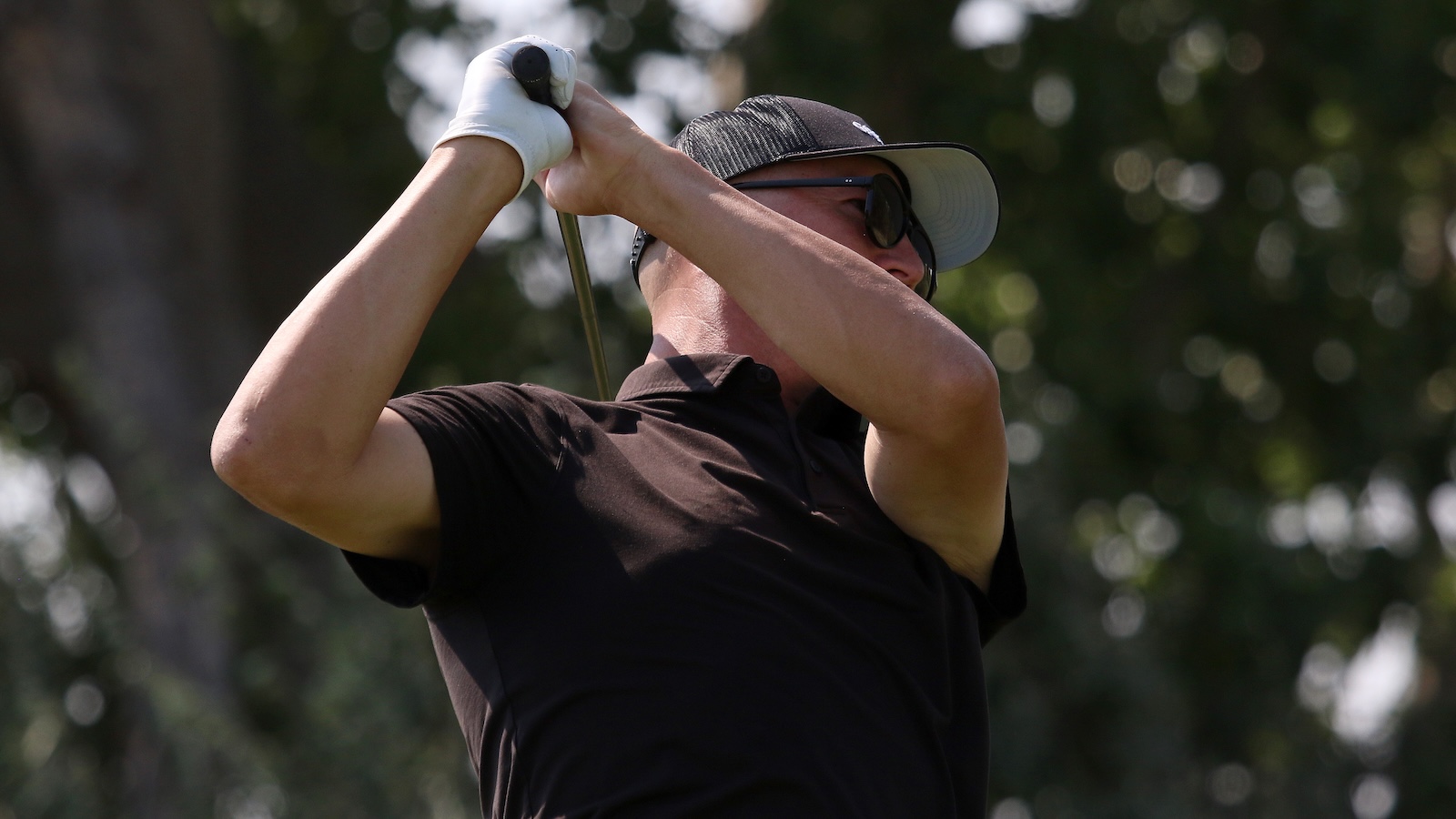 Golfer wearing a cap and sunglasses mid-swing on a sunny golf course, white-gloved hand gripping the club.