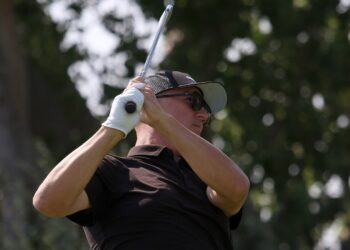 1K6A4196 | Press Pass Sports Male golfer in a black polo swinging a golf club, wearing a cap and sunglasses.