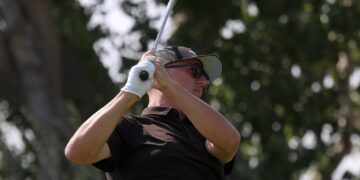 Male golfer in a black polo swinging a golf club, wearing a cap and sunglasses.