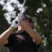 Male golfer in a black polo swinging a golf club, wearing a cap and sunglasses.